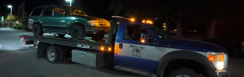 The truck at night in a lit lot, amber lights on, a green SUV on the bed